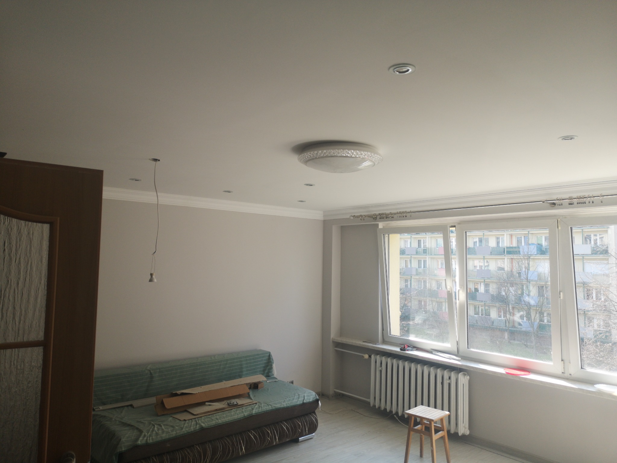 Pokój w trakcie odświeżania: świeżo pomalowane ściany w odcieniach szarości, dekoracyjny sufit z oświetleniem punktowym i wiszącym przewodem, okno z widokiem na budynek mieszkalny, zakryta sofa...