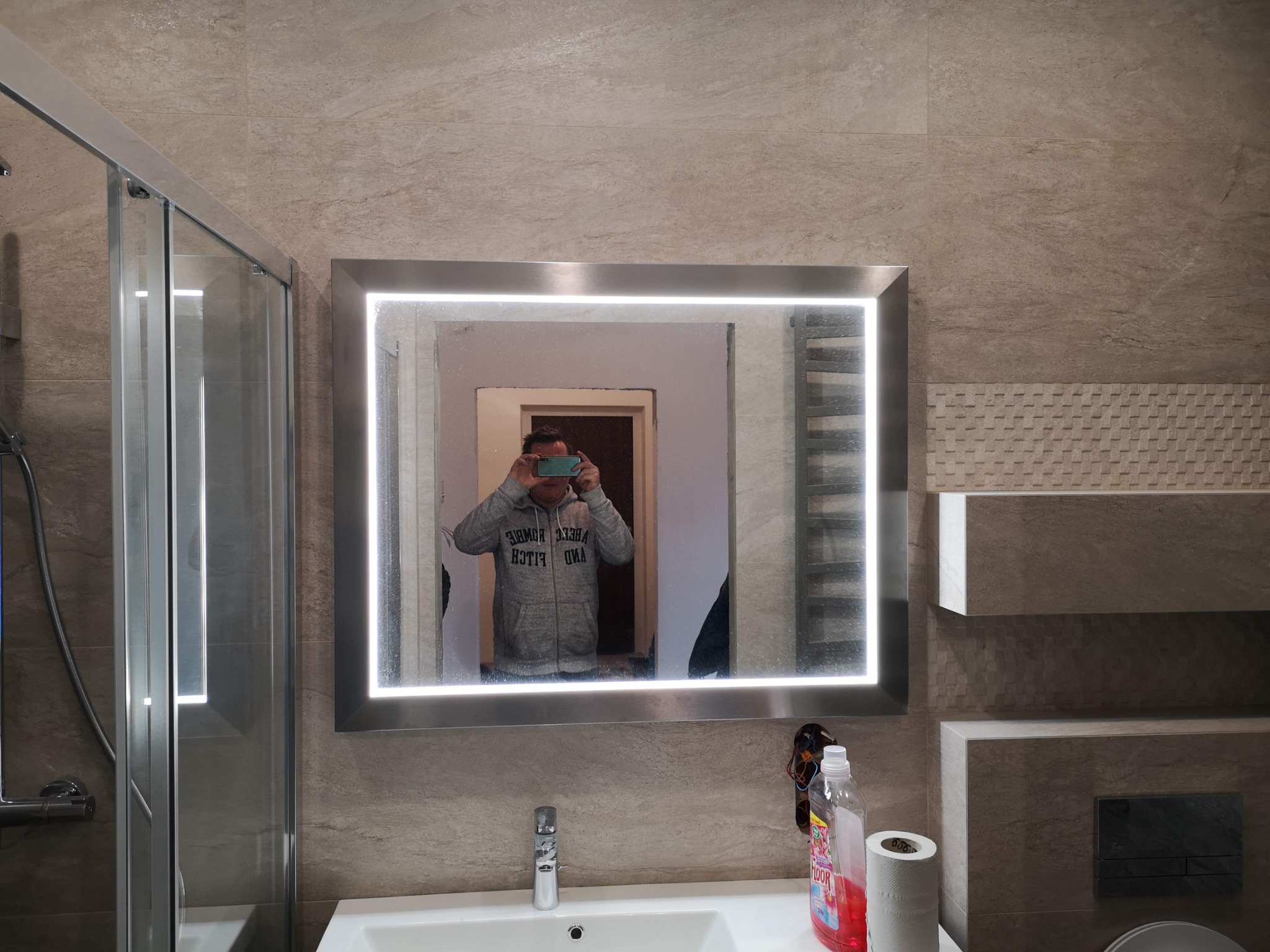 Nowoczesna łazienka z beżowymi płytkami, kabiną prysznicową i podświetlanym lustrem, w którym odbija się osoba robiąca zdjęcie telefonem.