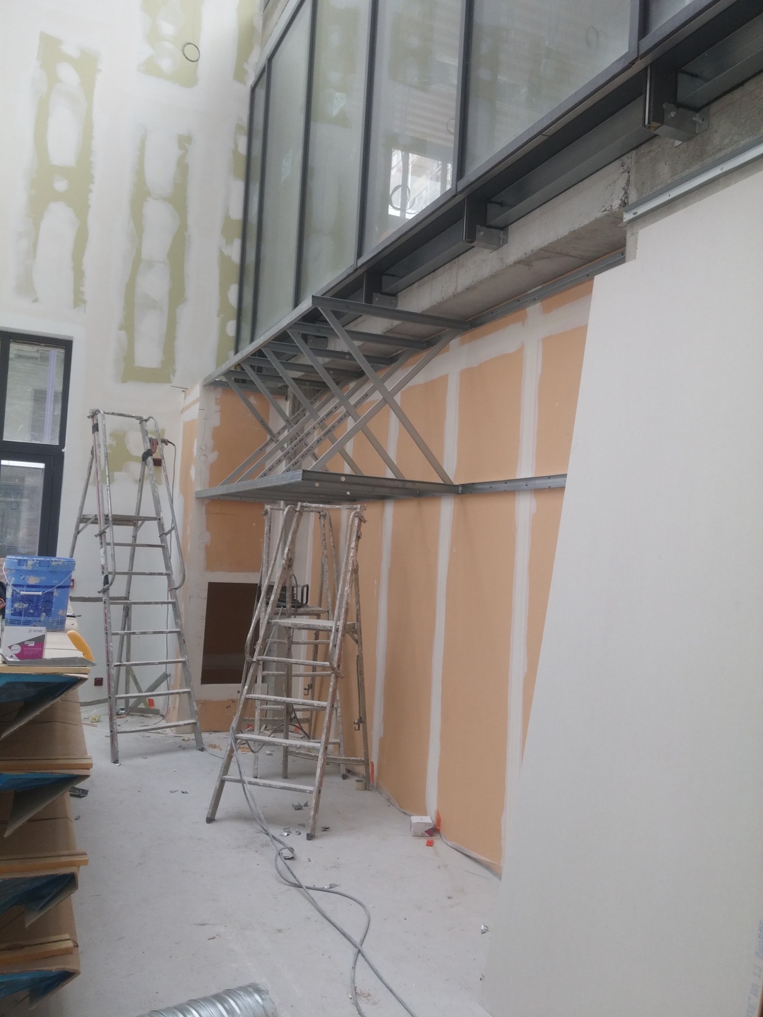 Wnętrze w trakcie prac z płytami gipsowo-kartonowymi, widoczne dwie drabiny, metalowa konstrukcja pod sufitem i częściowo zamontowane płyty na ścianach.
