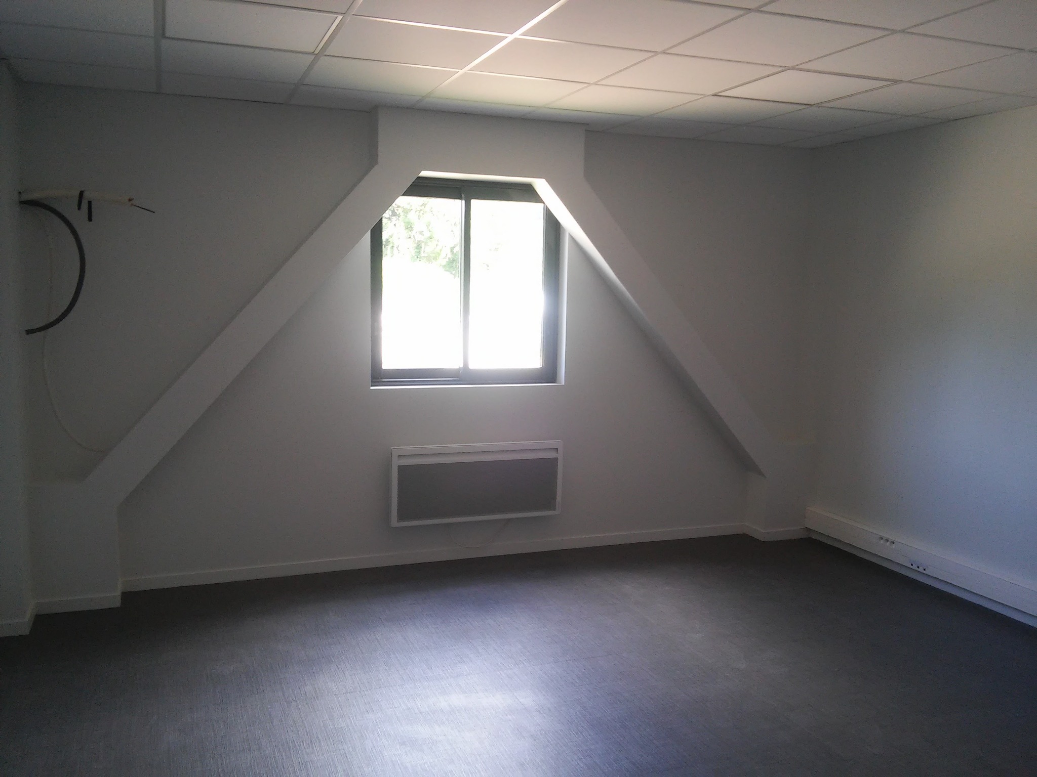 Puste poddasze z oknem w szczycie, szarą podłogą i białymi ścianami, widoczny grzejnik panelowy i listwy przypodłogowe.