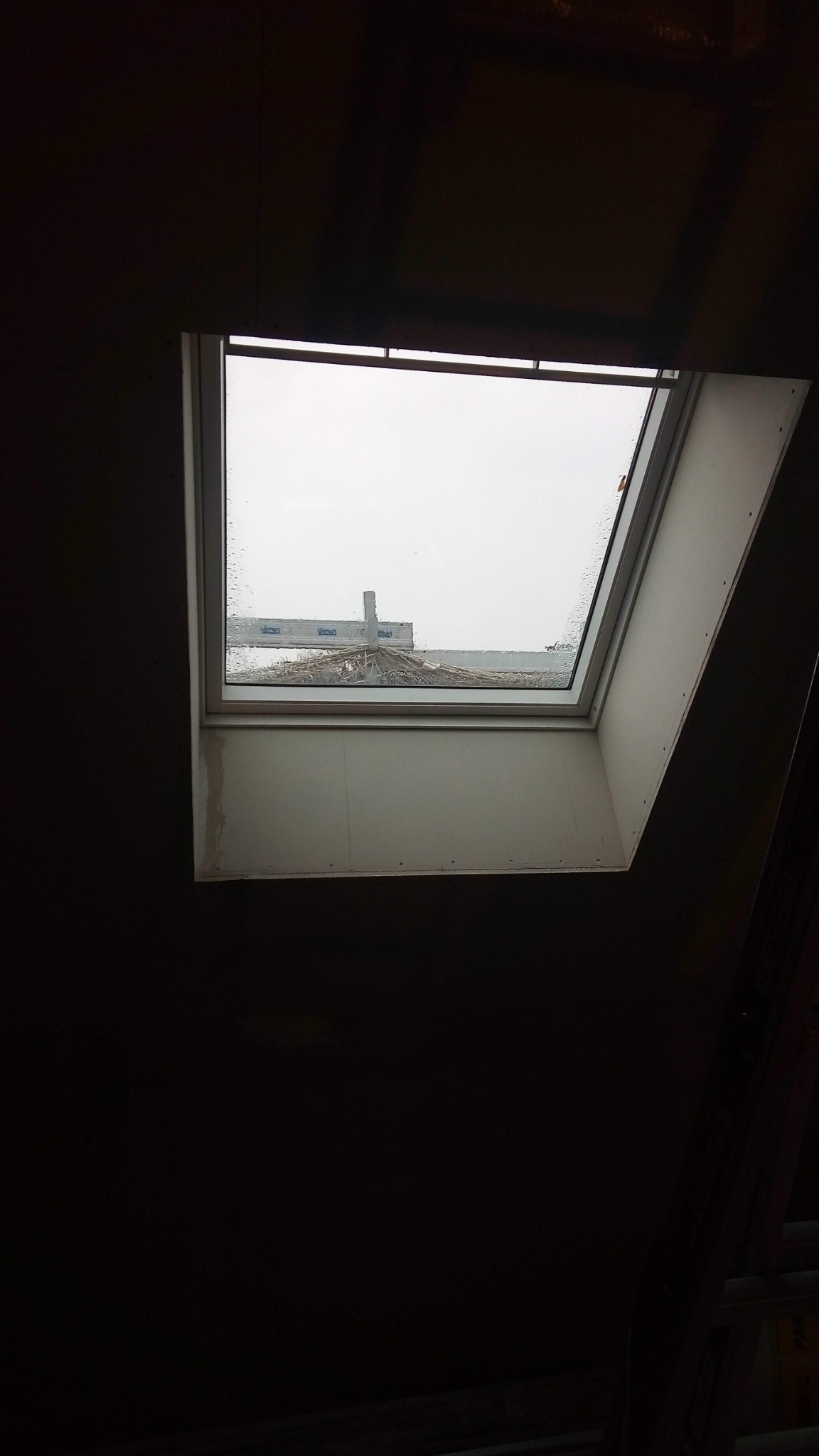 Wnętrze poddasza z zamontowanym oknem dachowym, widoczne krople deszczu na szybie i fragment dachu sąsiedniego budynku za oknem.