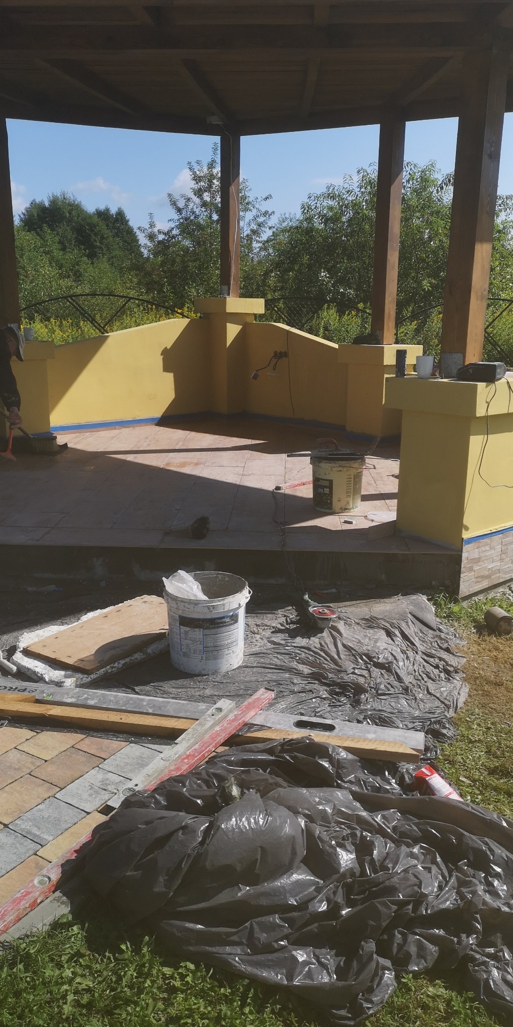 Budowa altany: żółte ściany, drewniane filary, płytki na podłodze, widoczne narzędzia i materiały budowlane w trakcie prac.