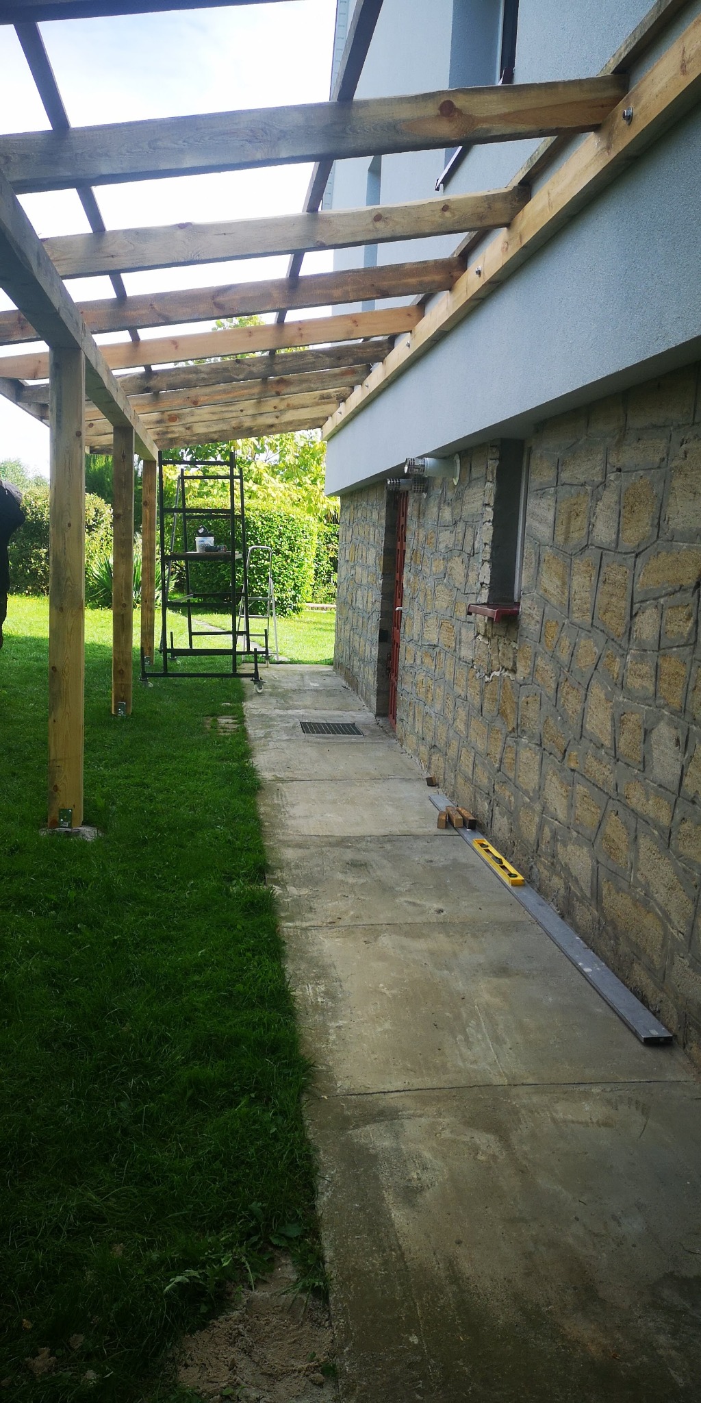 Drewniana konstrukcja zadaszenia tarasu w trakcie budowy, widoczna z zewnątrz, z elementami mocującymi do ściany budynku wykończonej kamieniem, obok trawnik i stojak na rośliny.