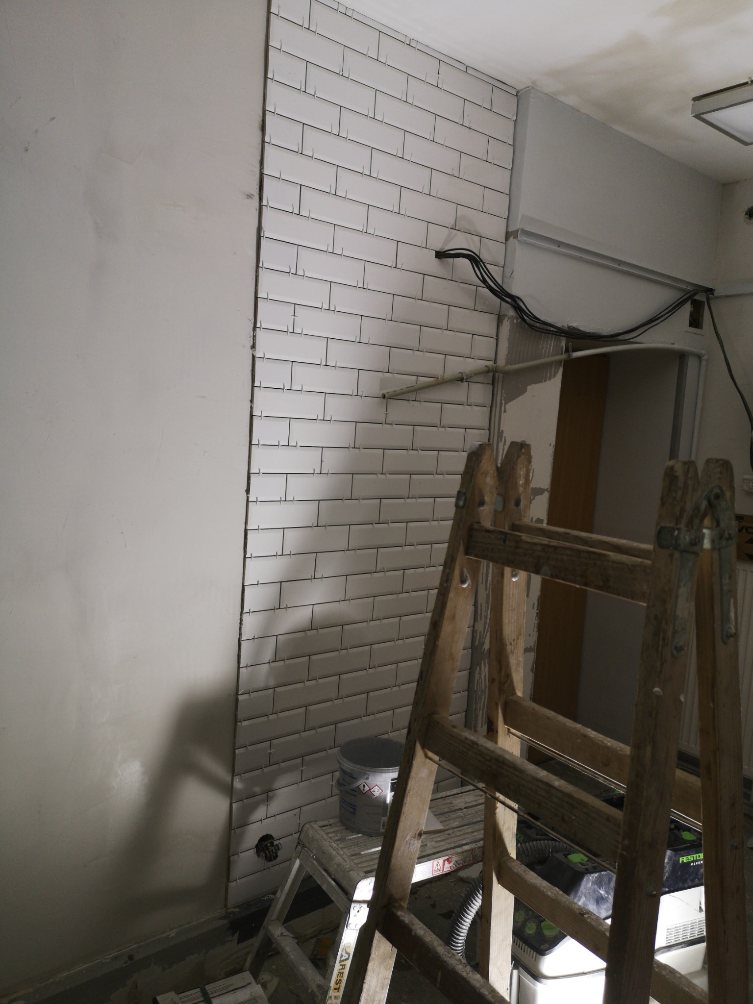 Fragment ściany z białymi, prostokątnymi płytkami w trakcie układania, widoczna drabina drewniana, wiadro z zaprawą i odkurzacz przemysłowy.
