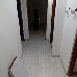 Malarz Pomorskie - Korytarz w trakcie prac w Gdańsku: widoczne ułożone panele podłogowe o jasnym, drewnopodobnym wzorze, karton po panelach przy ścianie, drabina w oddali oraz przygotowane gniazdka elektryczne.