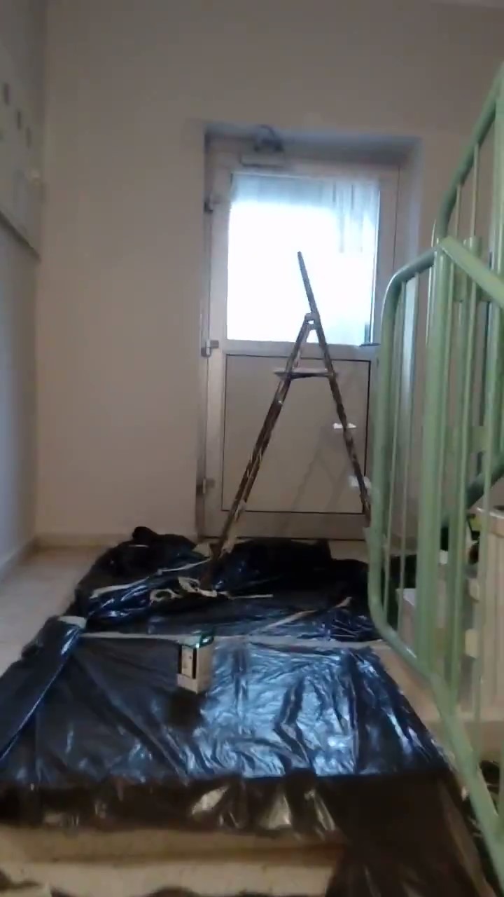 Przygotowanie do malowania klatki schodowej: podłoga zabezpieczona folią malarską, drabina oparta o ścianę przy drzwiach wejściowych, widoczna zielona balustrada schodów.