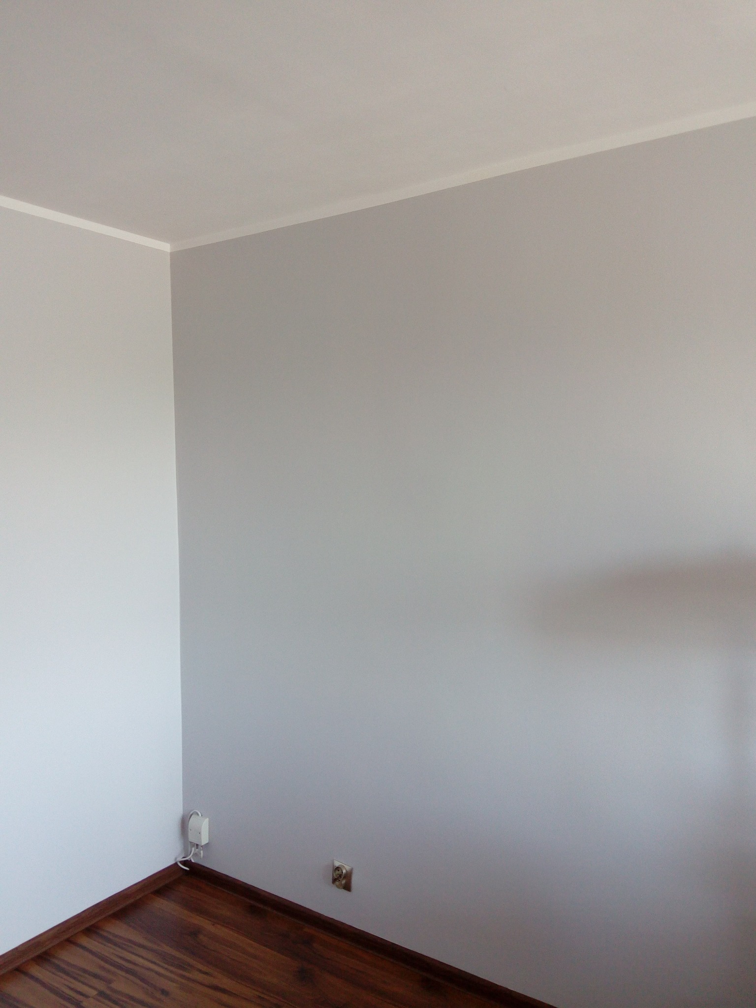 Pomieszczenie po malowaniu na szaro, widoczny fragment ściany z listwą przypodłogową w kolorze drewna, gniazdko elektryczne i biały kabel zasilający przy podłodze.