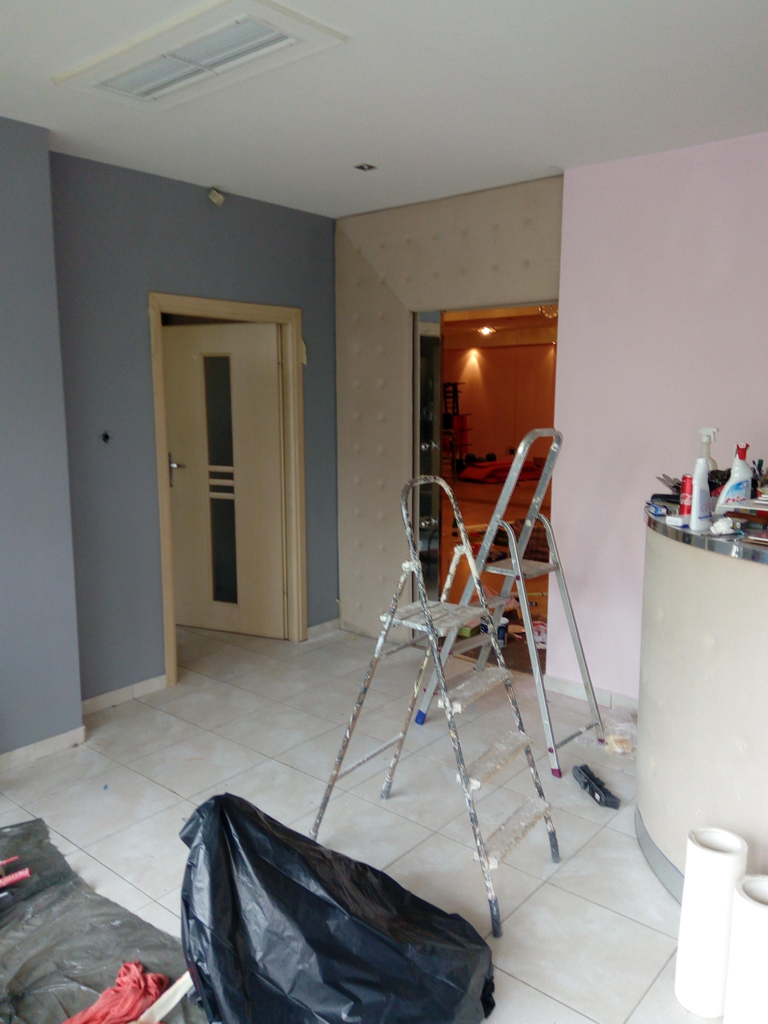 Pokój w trakcie malowania, ściany w odcieniach szarości i różu, drabina aluminiowa, zabezpieczona podłoga folią, widoczne narzędzia malarskie i puszki z farbą na blacie.