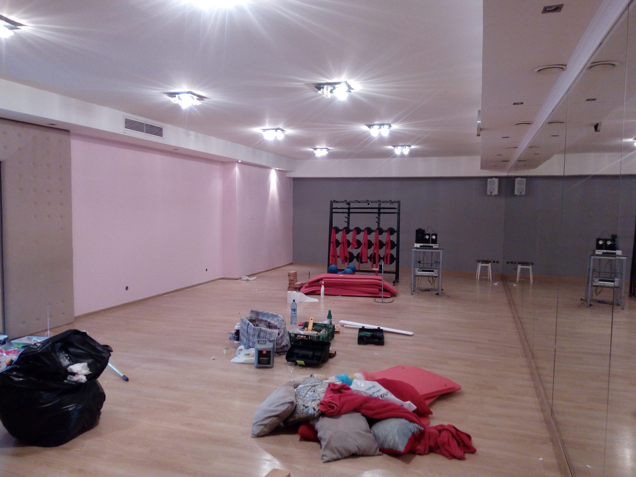 Wnętrze sali fitness w trakcie remontu: różowe i szare ściany, parkiet w jasnym odcieniu, rozrzucone materiały budowlane i worki na śmieci, widoczne lustra na ścianie.