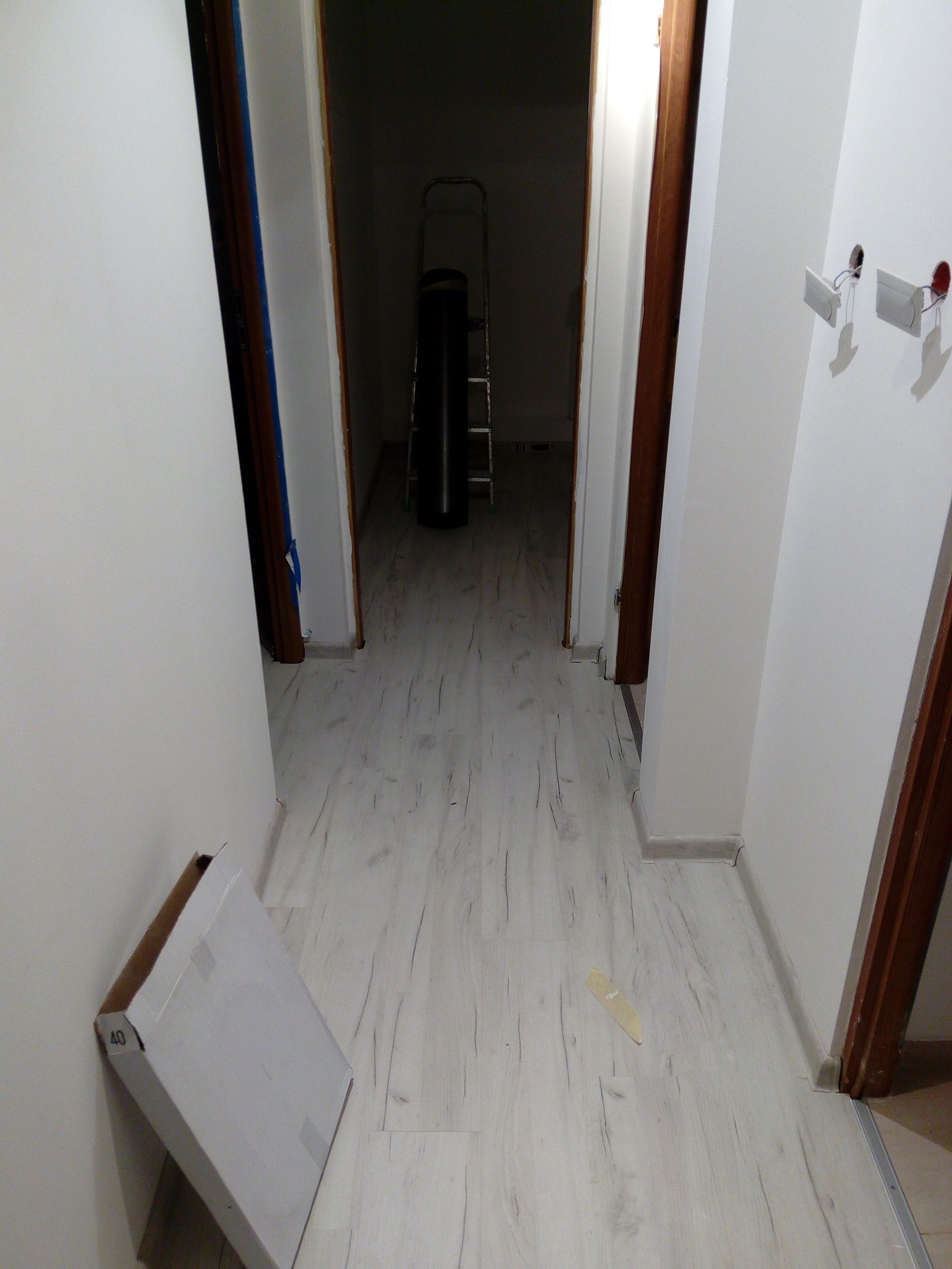 Korytarz w trakcie prac w Gdańsku: widoczne ułożone panele podłogowe o jasnym, drewnopodobnym wzorze, karton po panelach przy ścianie, drabina w oddali oraz przygotowane gniazdka elektryczne.