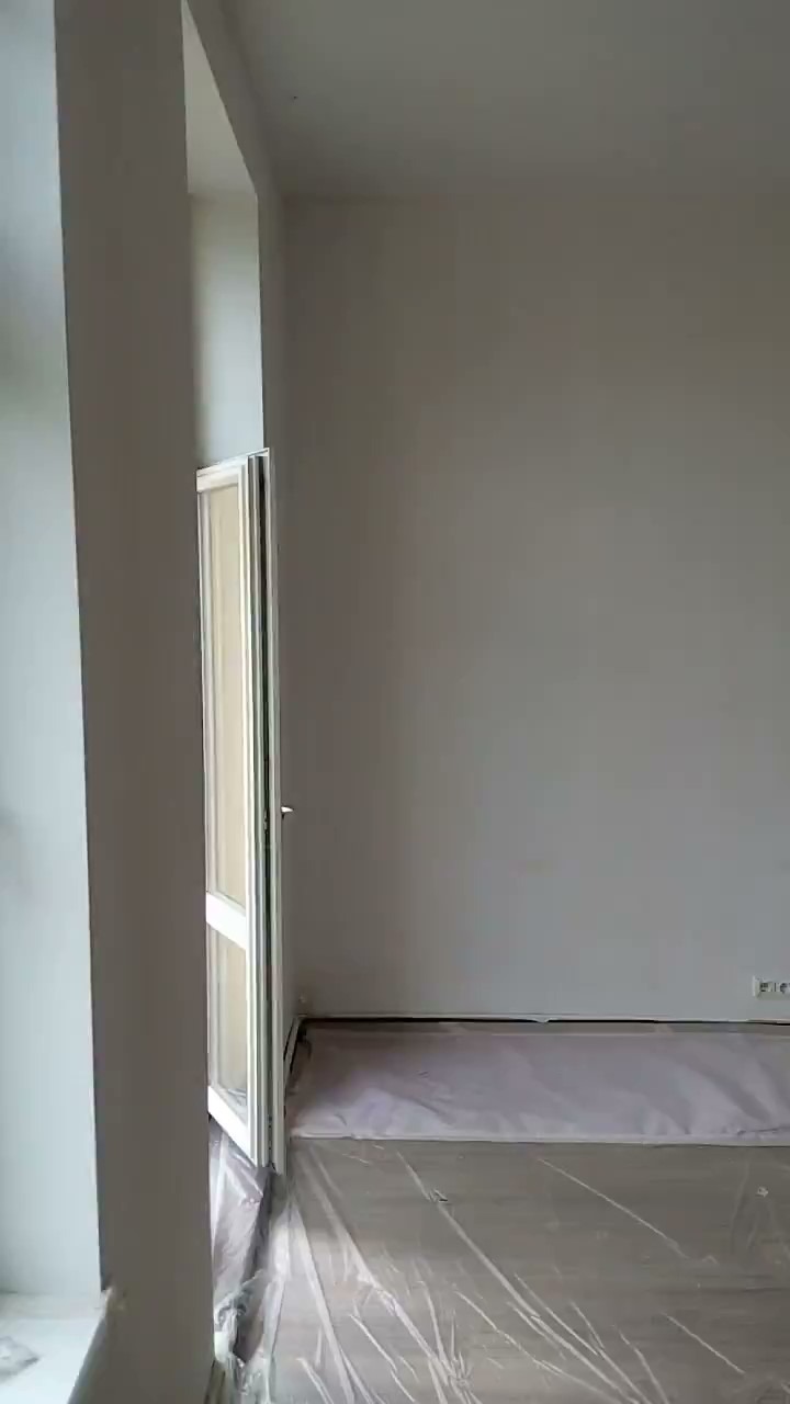 Świeżo pomalowane na szaro ściany w pokoju, podłoga zabezpieczona folią malarską, widoczne gniazdka elektryczne i otwarte okno.