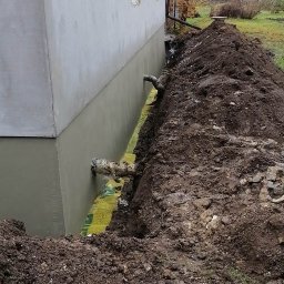 Usług budowlanych / praca/ wykończenia / itd - Fundament budynku po nałożeniu izolacji pionowej, widoczny wykop z żółtą folią izolacyjną i rurami odprowadzającymi wodę.