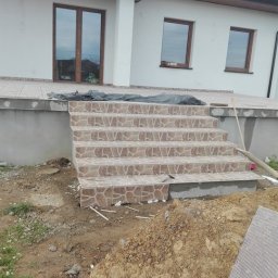 Usług budowlanych / praca/ wykończenia / itd - Wyłożone płytkami schody wejściowe do domu jednorodzinnego, widoczne prace wykończeniowe, niezagospodarowany teren wokół z widoczną pryzmą ziemi.