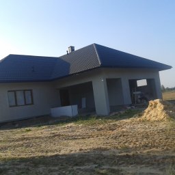 Usług budowlanych / praca/ wykończenia / itd - Niedokończony dom jednorodzinny z szarą elewacją i ciemnym dachem z blachodachówki, widoczny stos piasku i częściowo otwarty garaż z rusztowaniem w środku.