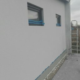 Usług budowlanych / praca/ wykończenia / itd - Boczny widok na świeżo pomalowaną białą elewację domu z dwoma oknami zabezpieczonymi niebieską taśmą malarską, widoczna drabina i ślady farby na chodniku.