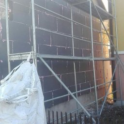 Usług budowlanych / praca/ wykończenia / itd - Ściana budynku w trakcie prac izolacyjnych, pokryta czarnymi płytami izolacyjnymi z widocznymi różowymi spoinami, otoczona metalowym rusztowaniem i workiem z materiałami budowlanymi.