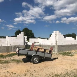 Usług budowlanych / praca/ wykończenia / itd - Wznoszenie ścian domu z białych bloczków betonu komórkowego, widoczna przyczepka z piaskiem na tle błękitnego nieba z chmurami.