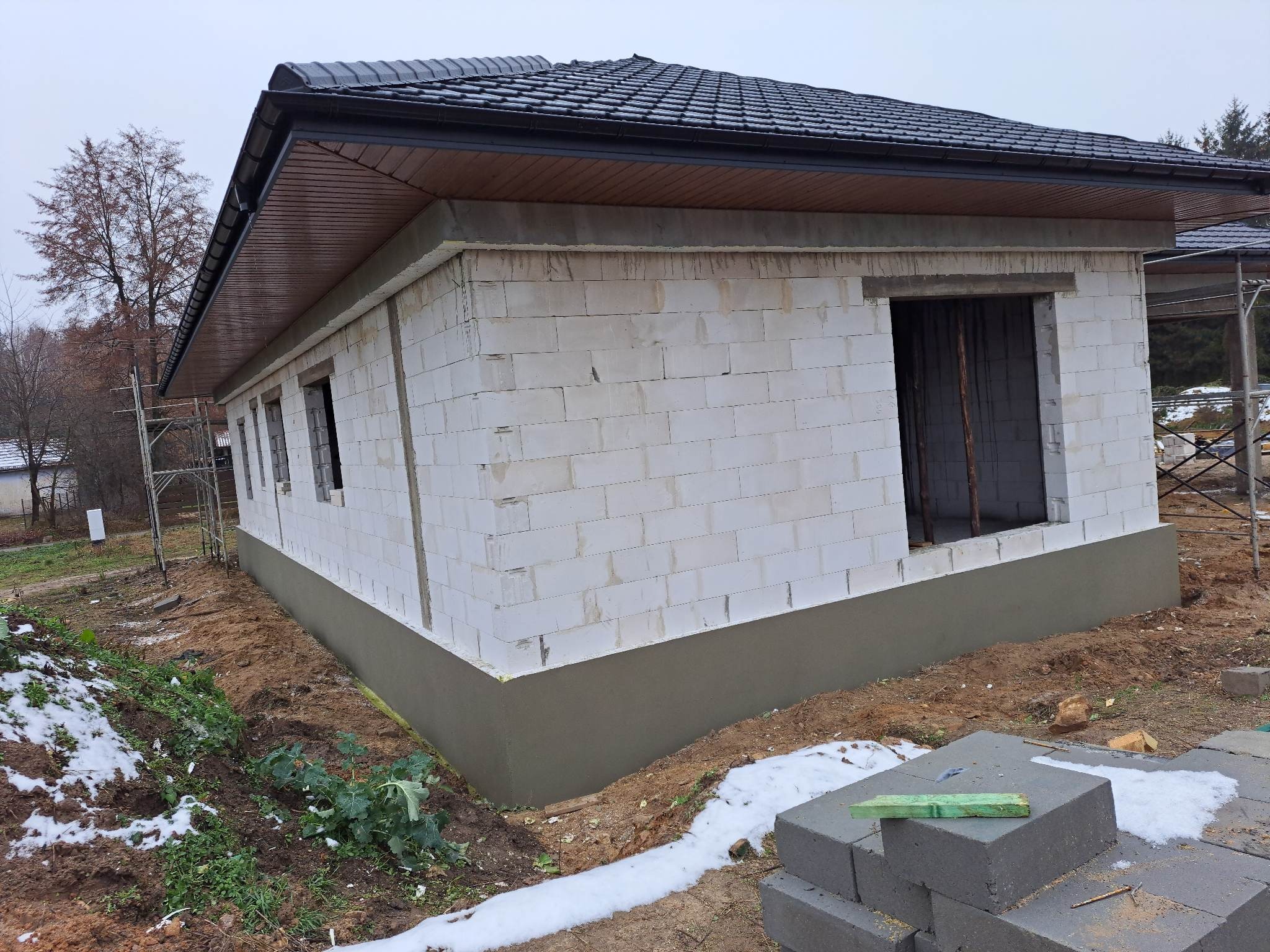 Budynek w trakcie budowy z białych bloczków, z widoczną izolacją fundamentów i ciemnym dachem, na zewnątrz resztki śniegu.