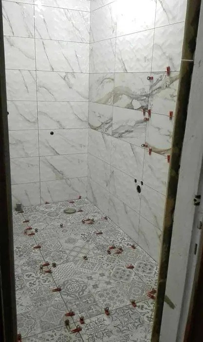 Łazienka w trakcie układania płytek podłogowych z wzorem patchworkowym i marmurowych płytek ściennych, widoczne krzyżyki dystansowe i otwory na instalacje.