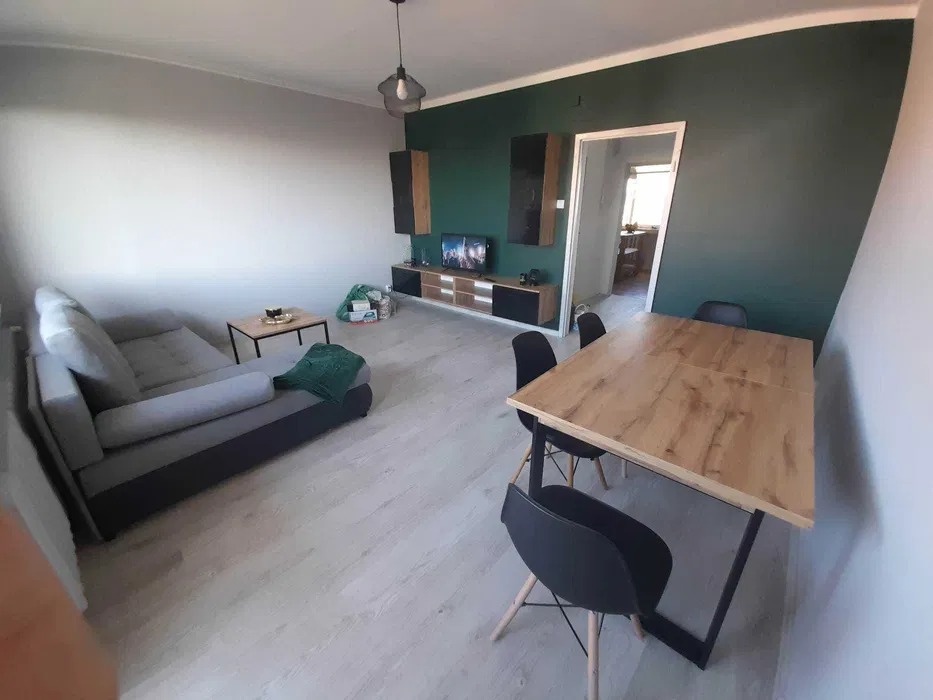 Jasne wnętrze pokoju dziennego z szarą sofą, drewnianym stołem z czarnymi krzesłami, ścianą w kolorze butelkowej zieleni i elementami dekoracyjnymi na ścianie.