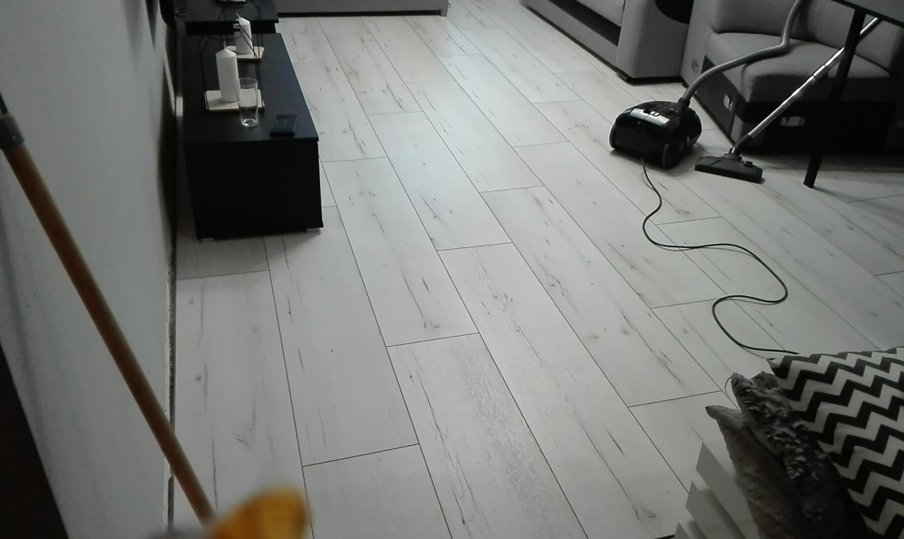 Jasne panele podłogowe po ułożeniu, z widocznym odkurzaczem i fragmentem mebli w tle, podczas sprzątania.