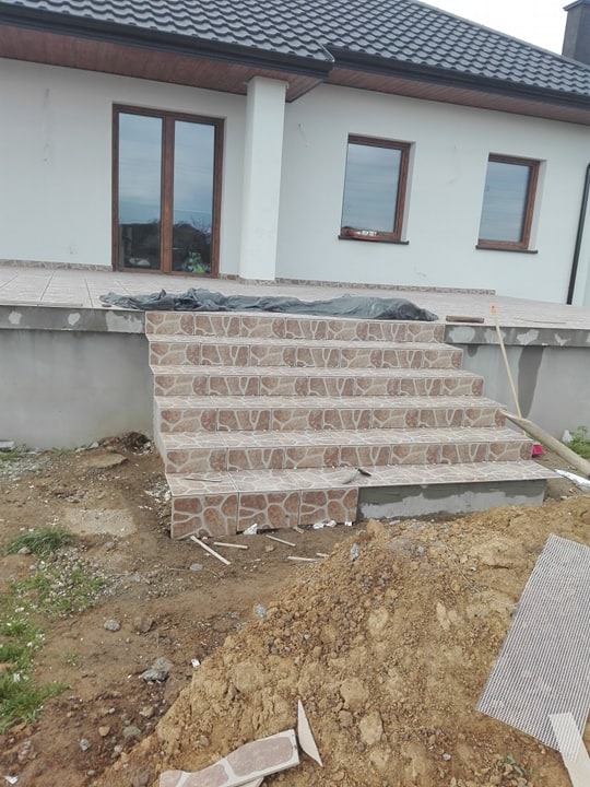 Wyłożone płytkami schody wejściowe do domu jednorodzinnego, widoczne prace wykończeniowe, niezagospodarowany teren wokół z widoczną pryzmą ziemi.