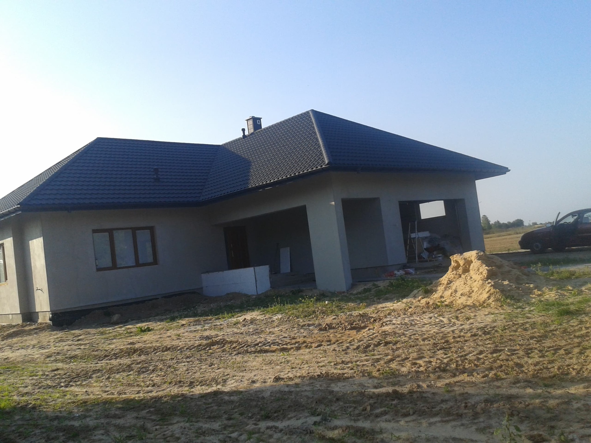 Niedokończony dom jednorodzinny z szarą elewacją i ciemnym dachem z blachodachówki, widoczny stos piasku i częściowo otwarty garaż z rusztowaniem w środku.