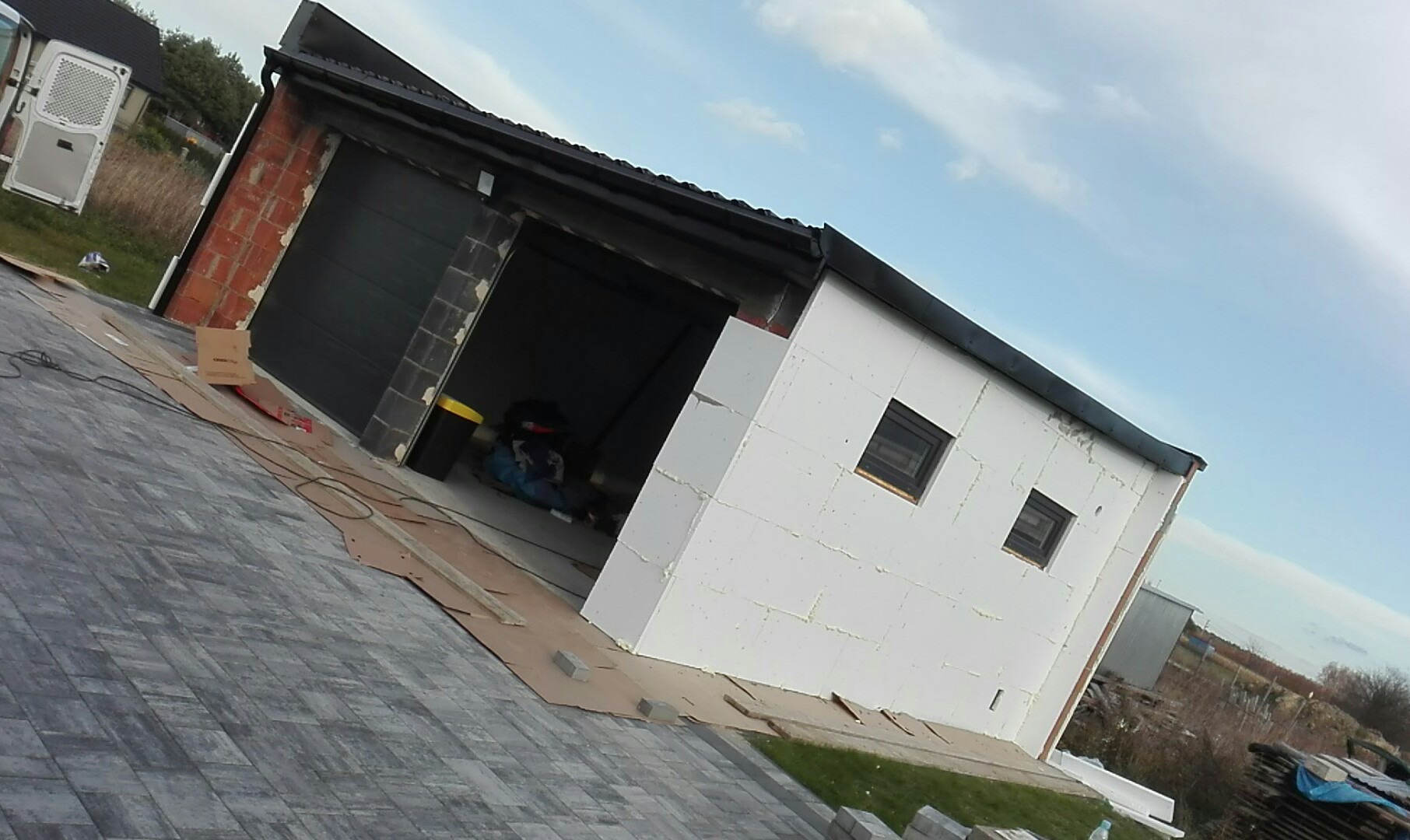 Garaż z czarną bramą segmentową, częściowo ocieplony białym styropianem, z widocznymi dwoma małymi oknami, na posesji z szarą kostką brukową i elementami wykończeniowymi na podłożu.