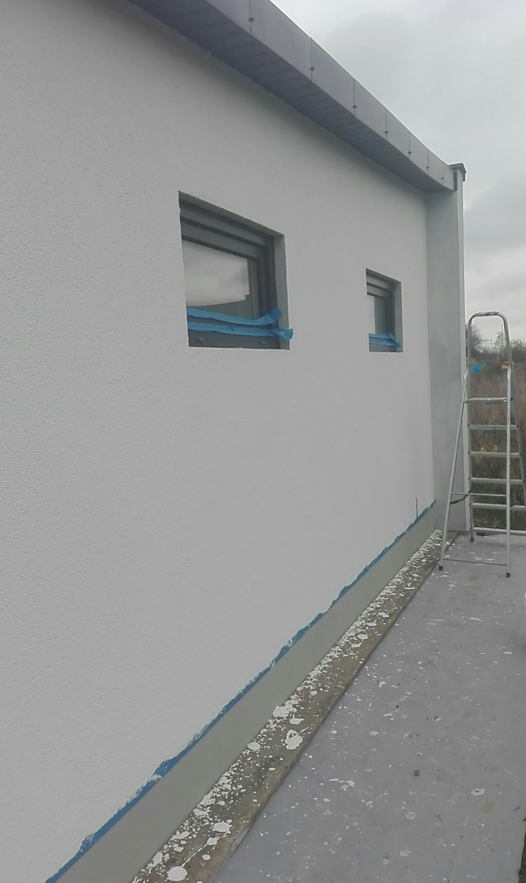 Boczny widok na świeżo pomalowaną białą elewację domu z dwoma oknami zabezpieczonymi niebieską taśmą malarską, widoczna drabina i ślady farby na chodniku.