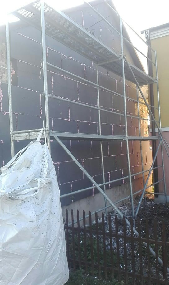 Ściana budynku w trakcie prac izolacyjnych, pokryta czarnymi płytami izolacyjnymi z widocznymi różowymi spoinami, otoczona metalowym rusztowaniem i workiem z materiałami budowlanymi.