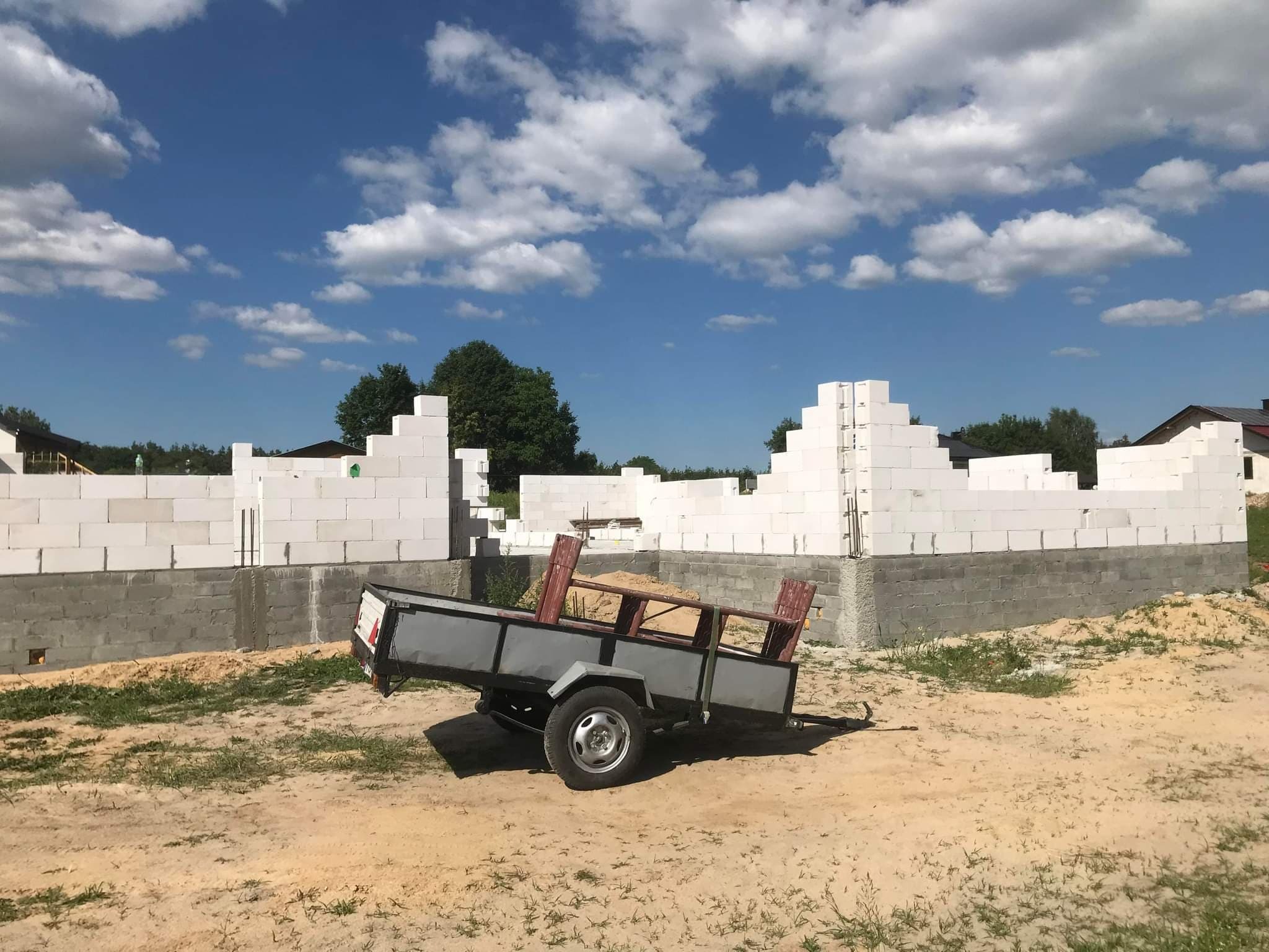 Wznoszenie ścian domu z białych bloczków betonu komórkowego, widoczna przyczepka z piaskiem na tle błękitnego nieba z chmurami.