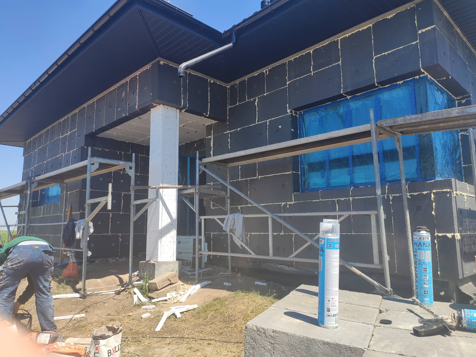 Dom jednorodzinny w trakcie prac izolacyjnych elewacji, widoczne czarne płyty styropianowe z wypełnionymi spoinami, rusztowania, zabezpieczone okna folią, puszki z pianą montażową i pracownik...