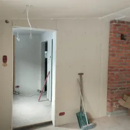 Wnętrze w trakcie remontu: ściany wyłożone płytami karton-gips, widoczny fragment komina z czerwonej cegły, narzędzia (szpadel i miotła) oparte o ścianę, instalacja elektryczna wyprowadzona ze ściany.