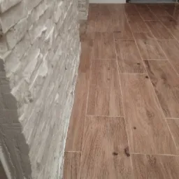 Ściana wykończona dekoracyjną, nieregularną cegłą w odcieniach bieli, sąsiadująca z podłogą wyłożoną podłużnymi płytkami imitującymi drewno w ciepłych, brązowych tonacjach.