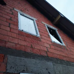 Surowa ściana z czerwonej cegły z dwoma zamontowanymi oknami z białymi ramami, jedno okno z szybą, drugie bez szyby, widok z dołu.