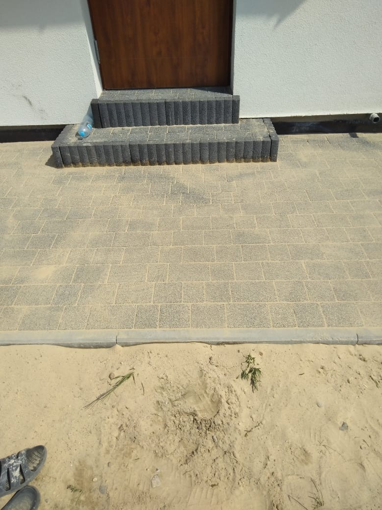 Ułożona kostka brukowa przed wejściem do domu z trzema stopniami, widoczna butelka wody i piasek obok.
