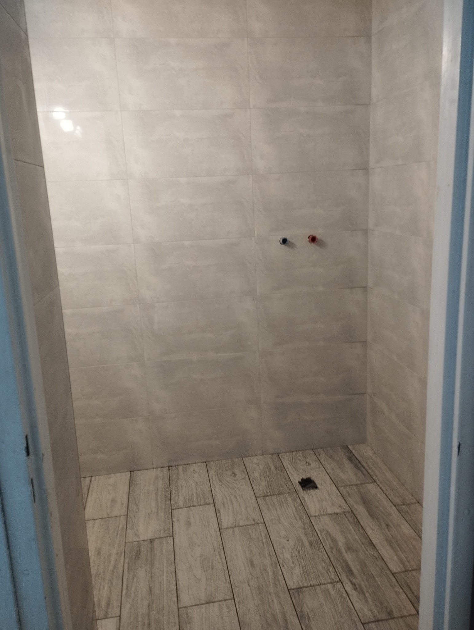 Wykończona kabina prysznicowa z jasnymi, imitującymi drewno płytkami na podłodze i szarymi, kwadratowymi płytkami na ścianach. Widoczne przyłącza wody z zaślepkami i odpływ liniowy.