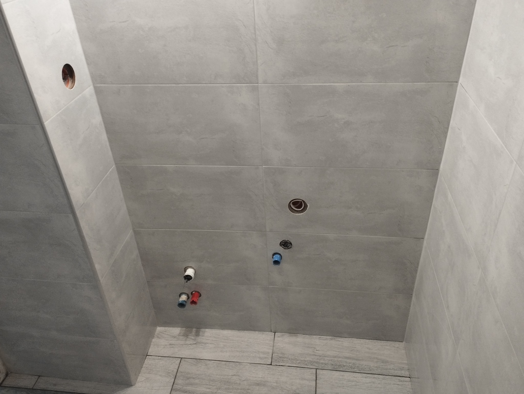 Szare, matowe płytki ceramiczne ułożone na ścianach łazienki, z widocznymi otworami na instalacje wodne i kanalizacyjne, w trakcie prac wykończeniowych.