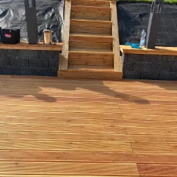 Nowa drewniana deska tarasowa z ryflowaną powierzchnią i schodami, widoczna skrzynka narzędziowa Makita, puszka z pędzlem i butelka wody na murku z bloczków betonowych.