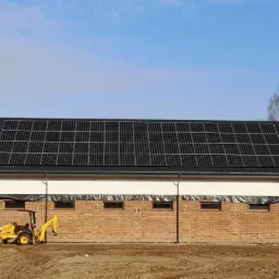 Budynek z cegły z panelami słonecznymi na dachu, żółta mini-ładowarka stoi przed budynkiem na placu budowy.