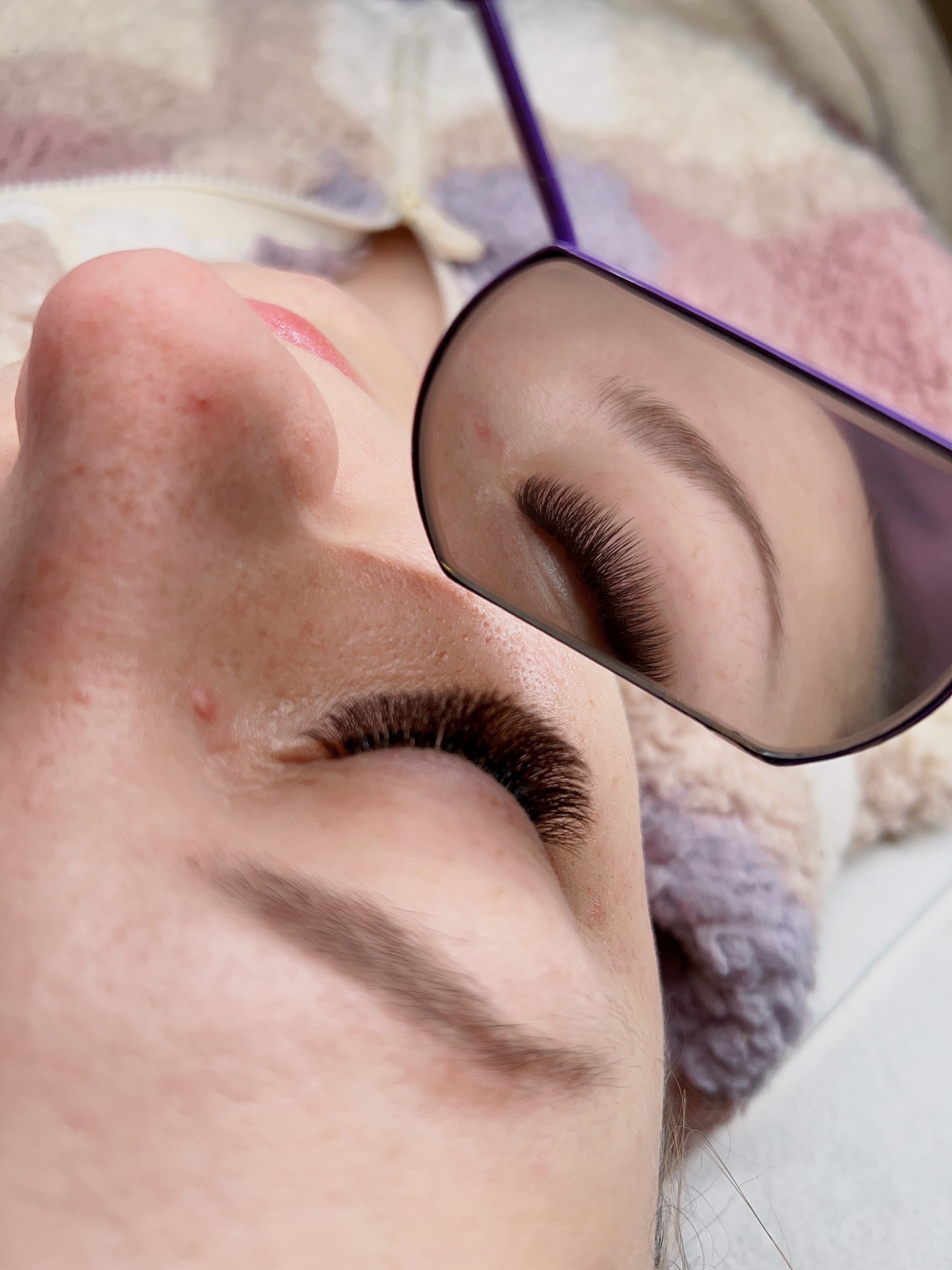 Zbliżenie na oko po zabiegu przedłużania rzęs, widoczne w lustrze, z delikatnym makijażem ust i fragmentem wzorzystej bluzy w tle.