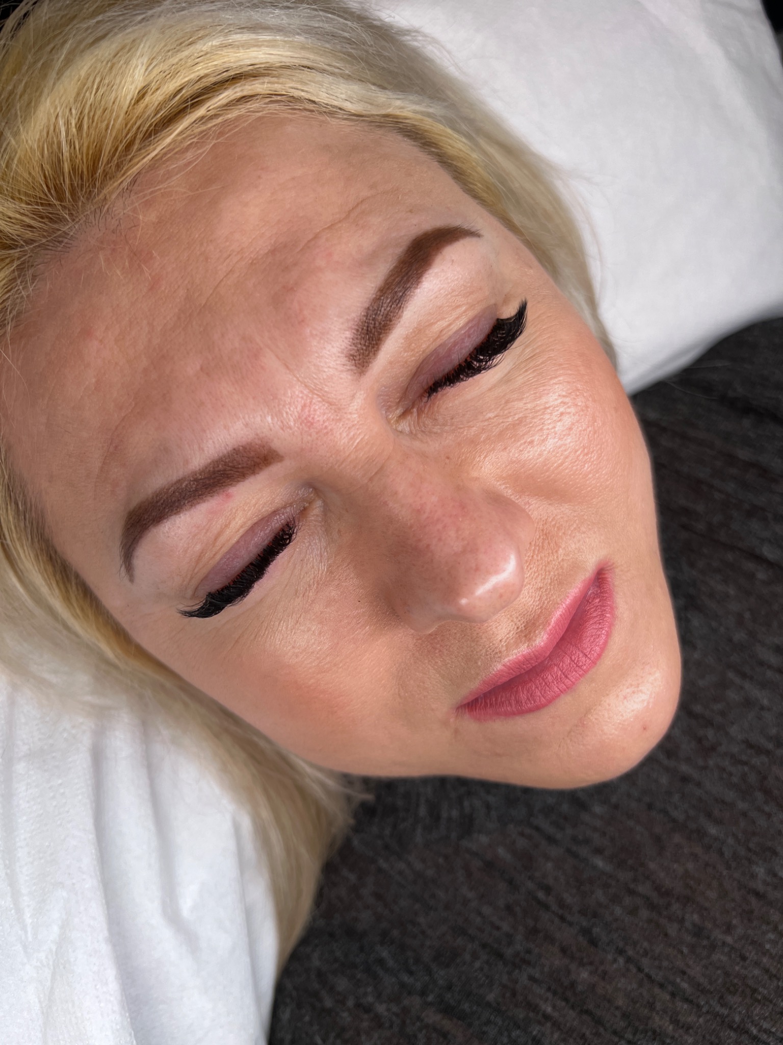 Kobieta z zamkniętymi oczami leżąca na białej poduszce po zabiegu makijażu permanentnego brwi i przedłużania rzęs, widoczne precyzyjnie wykonane brwi i długie, czarne rzęsy.