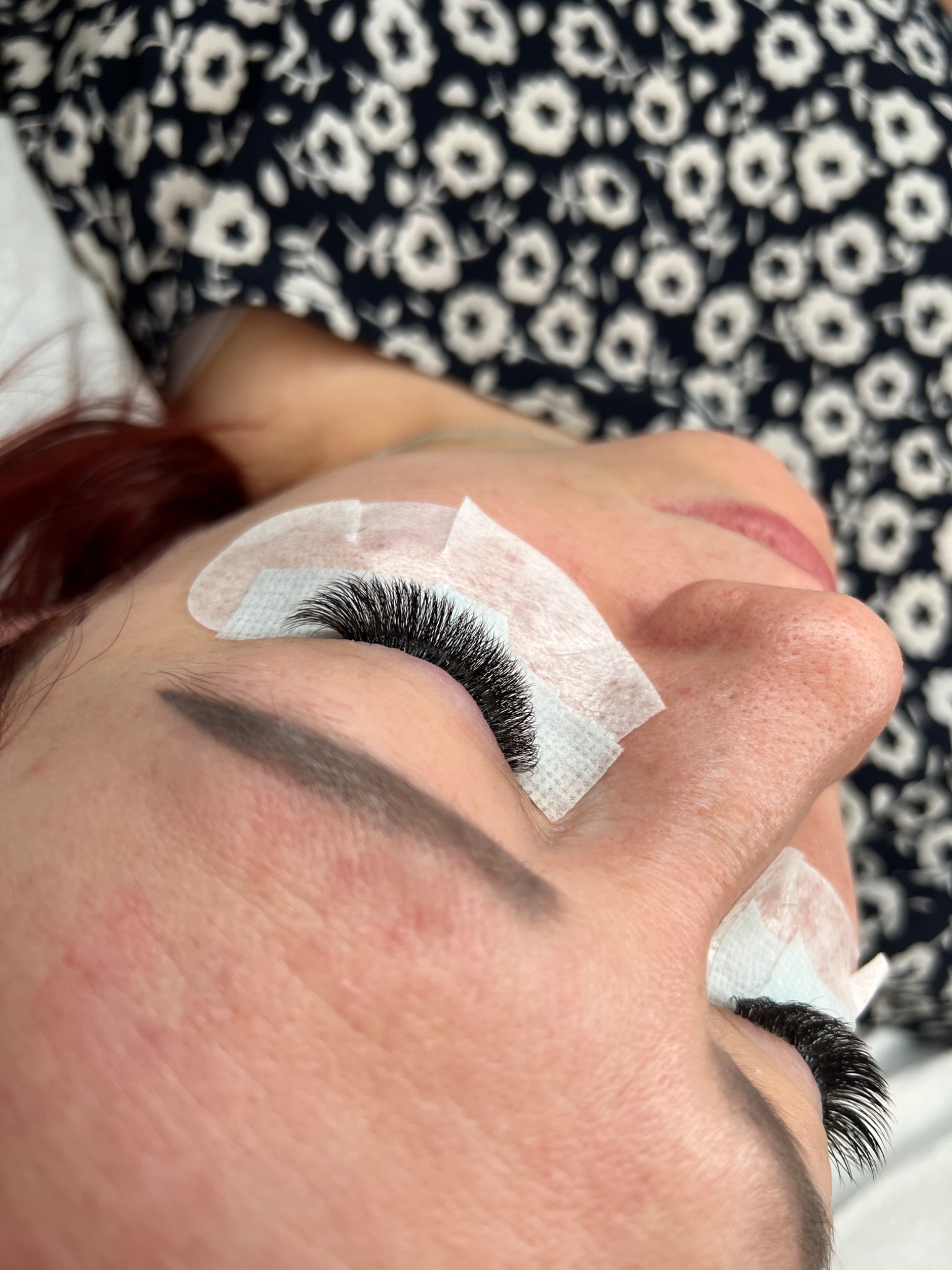 Kobieta z czerwonymi włosami leżąca na plecach z założonymi płatkami pod oczami i świeżo przedłużonymi, gęstymi rzęsami w stylu Russian Volume.