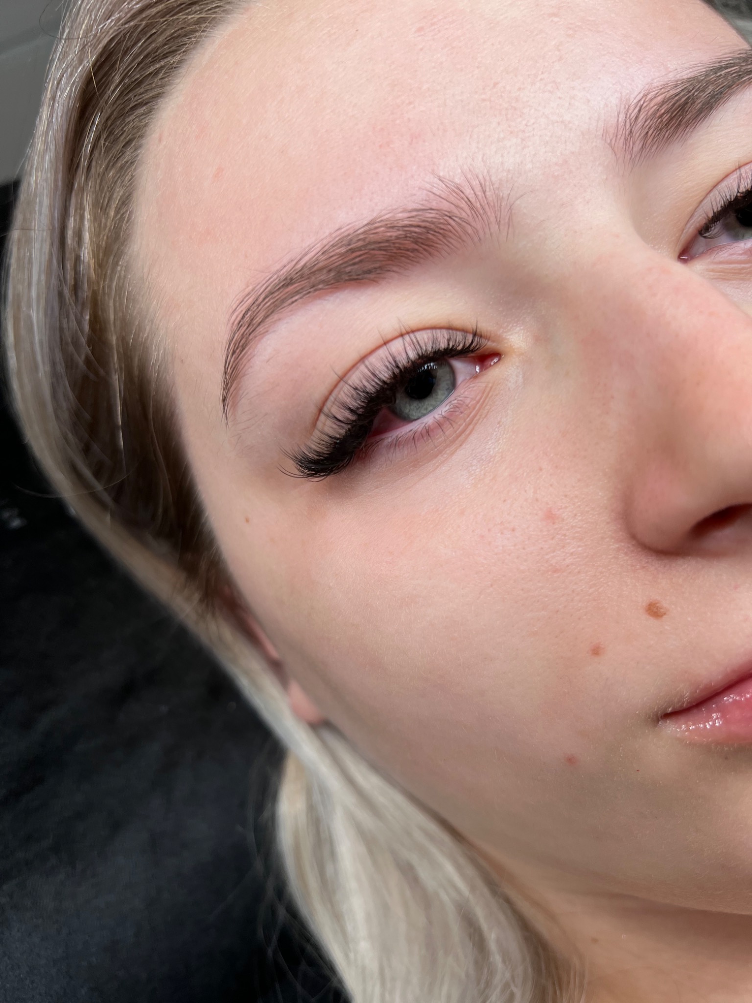 Zbliżenie na oko kobiety z profesjonalnie przedłużonymi rzęsami, widoczne naturalne brwi po zabiegu laminacji i regulacji, delikatny makijaż ust.