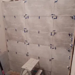 Układanie szarych płytek na ścianie w Łodzi, z użyciem niebieskich krzyżyków dystansowych i drabiny, w trakcie remontu łazienki.