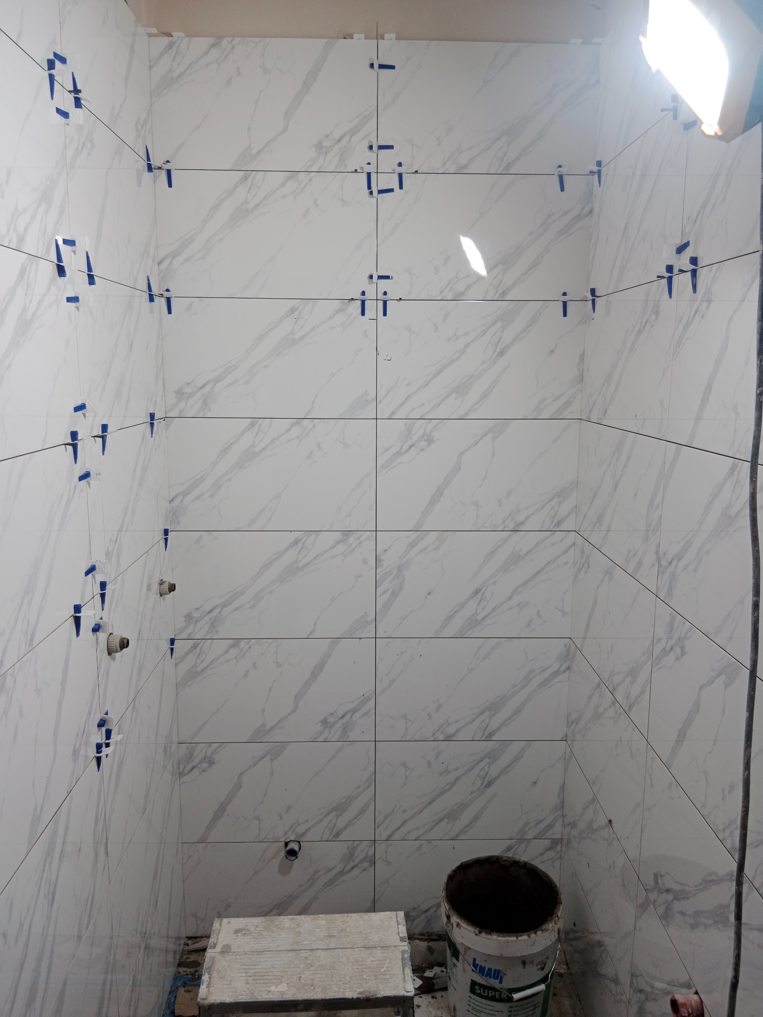 Ściany łazienki w trakcie układania białych płytek imitujących marmur, z użyciem poziomicy laserowej i klipsów poziomujących, widoczny kubeł z zaprawą klejową.
