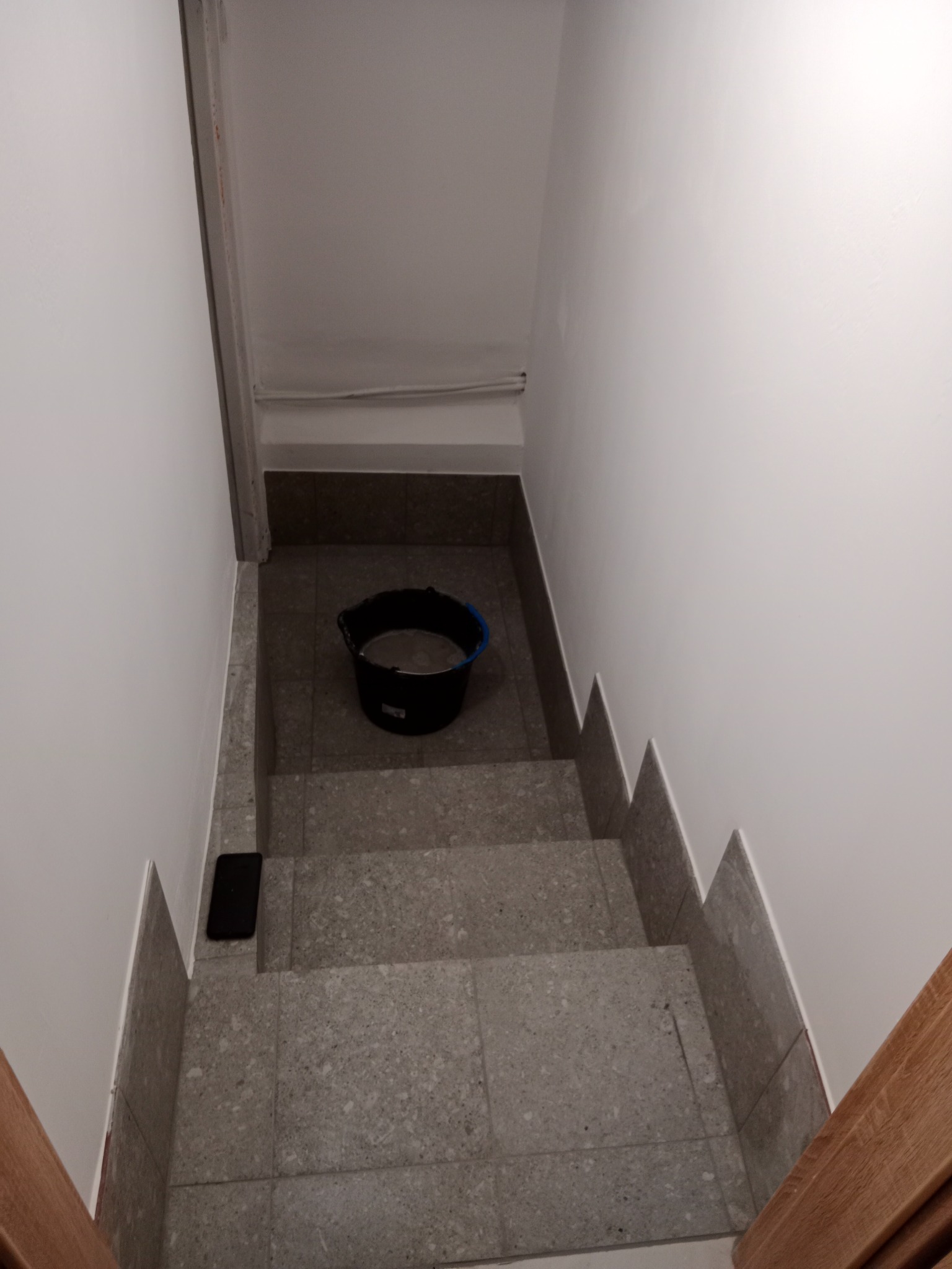 Schody wyłożone szarą terakotą z wiadrem na środku i telefonem leżącym na stopniu, ściany pomalowane na biało, widok z góry.