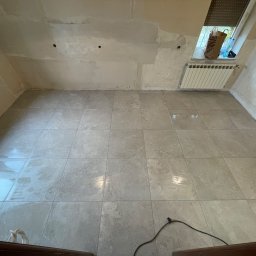 MARKDOM Usługi remontowo-wykończeniowe - Świeżo ułożona, jasnoszara podłoga z płytek w pomieszczeniu w trakcie remontu, ściany częściowo otynkowane, widoczne otwory w ścianie, grzejnik pod oknem ze skosami, kabel elektryczny na podłodze.