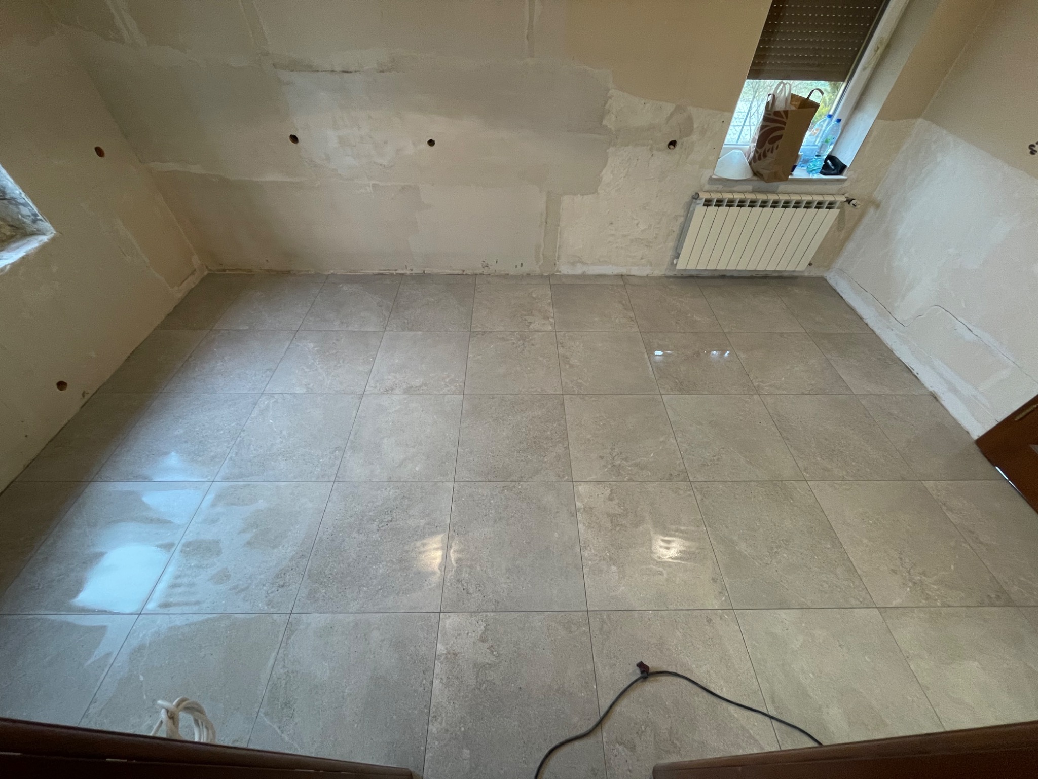 Świeżo ułożona, jasnoszara podłoga z płytek w pomieszczeniu w trakcie remontu, ściany częściowo otynkowane, widoczne otwory w ścianie, grzejnik pod oknem ze skosami, kabel elektryczny na podłodze.