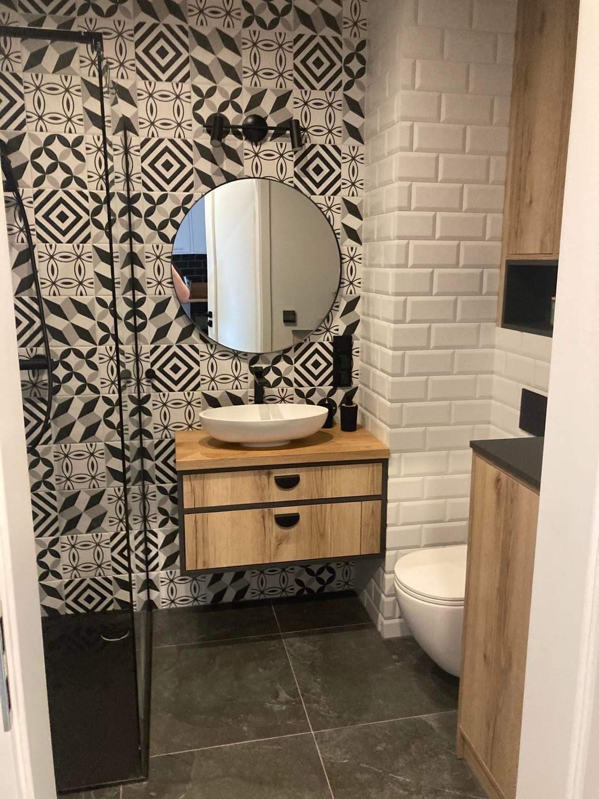 Nowoczesna łazienka z umywalką nablatową, szafką z drewnianym fornirem, czarną armaturą i geometrycznymi płytkami na ścianie w strefie prysznica.